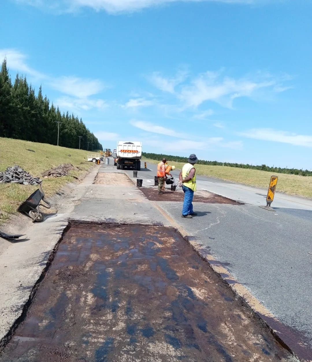 A rectangular section of road surfacing cut out and coated with black tack coat, crew working beyond.